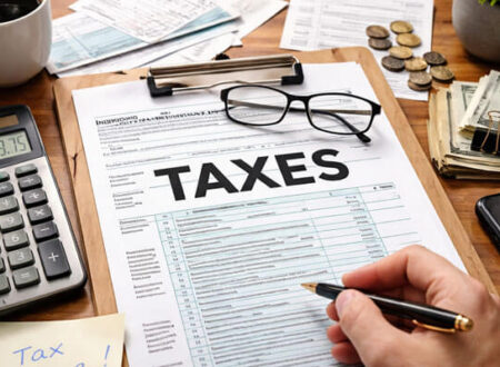 A close-up shot of a wooden desk featuring a U.S. Individual Income Tax Form 1040 with the word 'TAXES' prominently displayed. The scene includes a calculator showing $2,659.75, a cup of coffee, organized paperwork, a pair of glasses, and a hand holding a pen, ready to fill out the form. Nearby, dollar bills, coins, and a small potted plant add to the tax preparation ambiance.