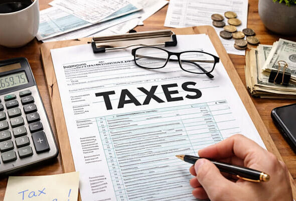 A close-up shot of a wooden desk featuring a U.S. Individual Income Tax Form 1040 with the word 'TAXES' prominently displayed. The scene includes a calculator showing $2,659.75, a cup of coffee, organized paperwork, a pair of glasses, and a hand holding a pen, ready to fill out the form. Nearby, dollar bills, coins, and a small potted plant add to the tax preparation ambiance.