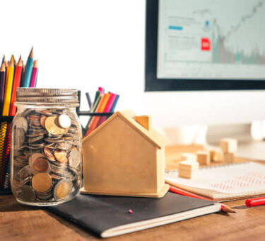 How to start investing for beginners concept with coins in a glass jar, small house model, financial notebook, and stock market chart on computer screen in a realistic workspace setup.