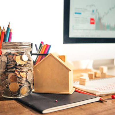 How to start investing for beginners concept with coins in a glass jar, small house model, financial notebook, and stock market chart on computer screen in a realistic workspace setup.
