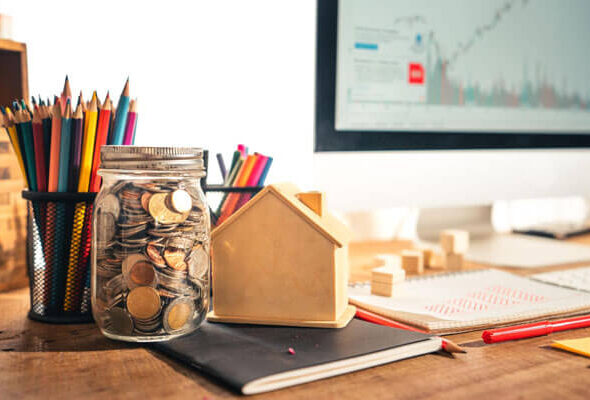How to start investing for beginners concept with coins in a glass jar, small house model, financial notebook, and stock market chart on computer screen in a realistic workspace setup.