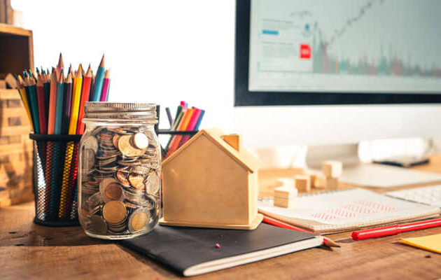 How to start investing for beginners concept with coins in a glass jar, small house model, financial notebook, and stock market chart on computer screen in a realistic workspace setup.