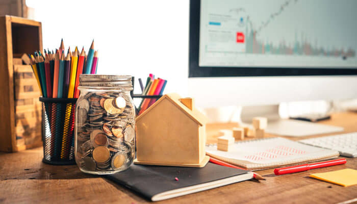 How to start investing for beginners concept with coins in a glass jar, small house model, financial notebook, and stock market chart on computer screen in a realistic workspace setup.